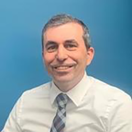 Man with graying hair, wearing a white shirt and blue-striped tie, smiling against a blue background.