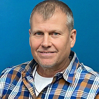 Man with gray hair and blue eyes smiles, wearing a plaid shirt and white undershirt against a blue backdrop.