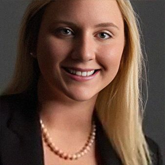 Woman with blonde hair smiles, wearing a pearl necklace and blazer, against a dark background.