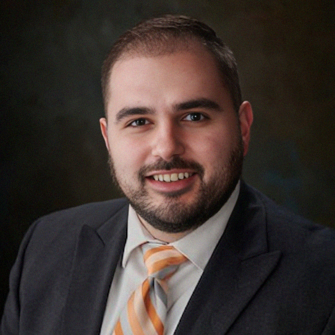 Man in a suit, smiling, with a patterned tie. Dark background.