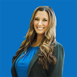 Woman with long brown hair, smiling, wearing a blue top and gray blazer, against a blue background.