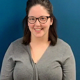 Woman with glasses smiles, wearing a gray sweater, in front of a blue wall.