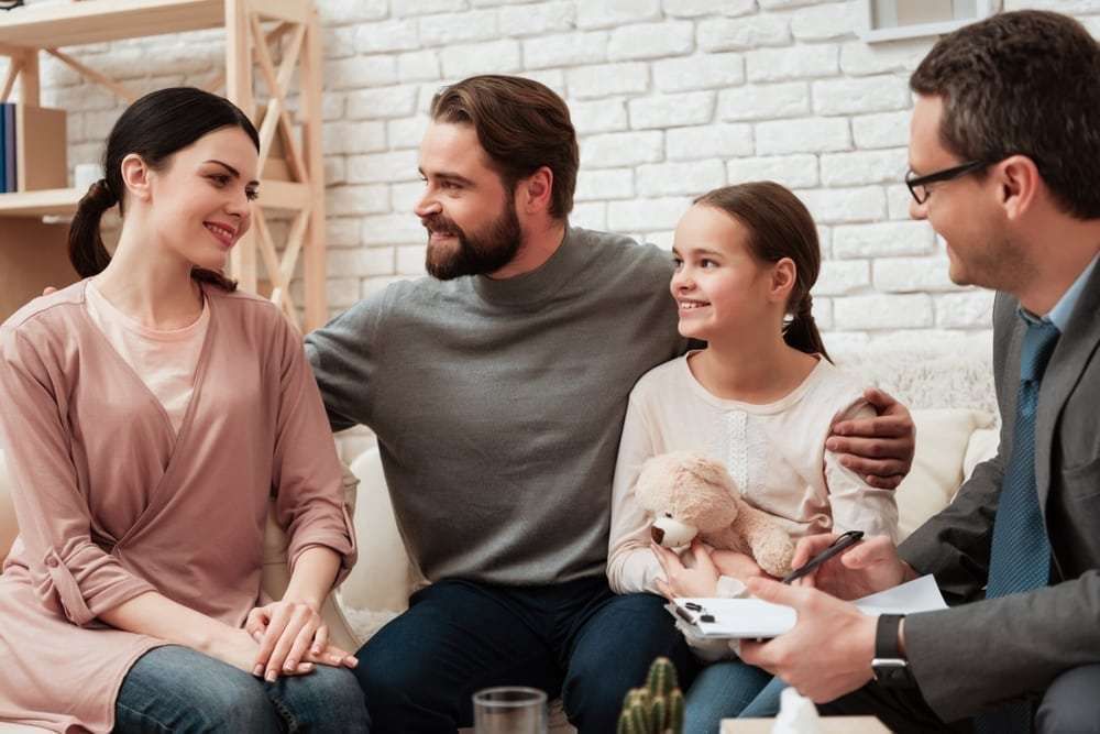 Family with a therapist, smiling, seated on a couch in a room with a window and a bookshelf.