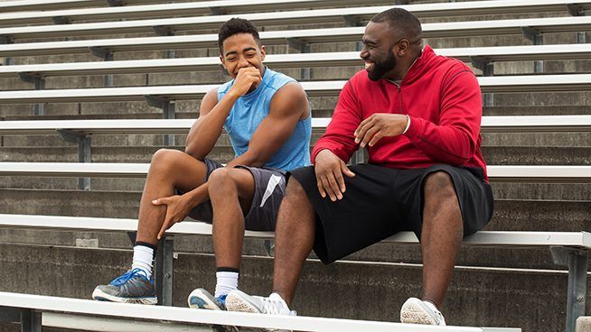 Two men, likely a coach and athlete, laugh while sitting on bleachers at a sports field.