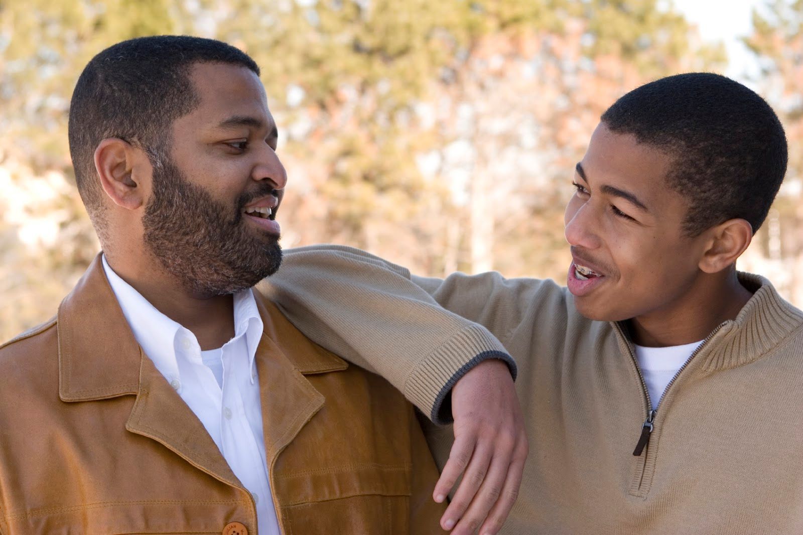 Father with a beard and son talking, outdoors. The son has arm on father's shoulder; both smiling.