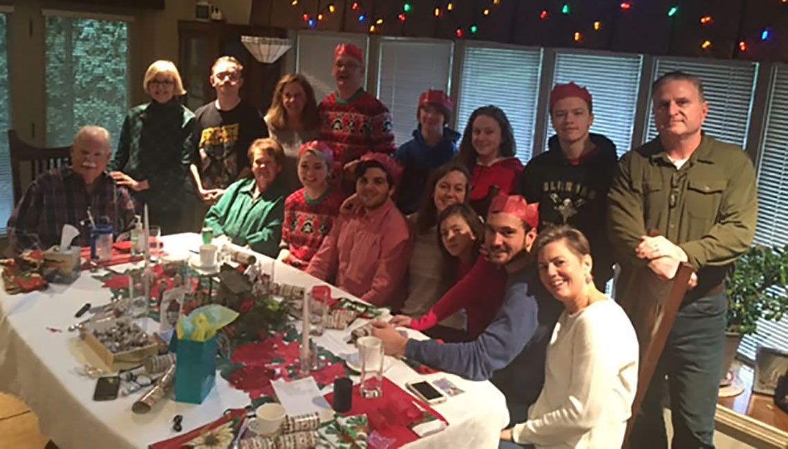Large family gathered at a table, many wearing festive hats. Christmas celebration in a home with string lights.