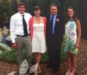 Family of four poses outdoors. A woman wears a white dress and pink sweater, a man in a suit, and two teens.