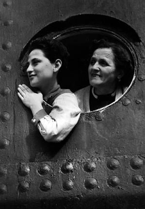 A young person and older woman look out of a round window, both smiling, possibly on a ship.