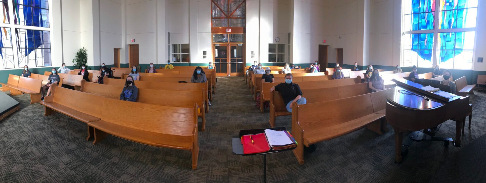 Church interior with wooden pews, stained glass windows, and people wearing masks.