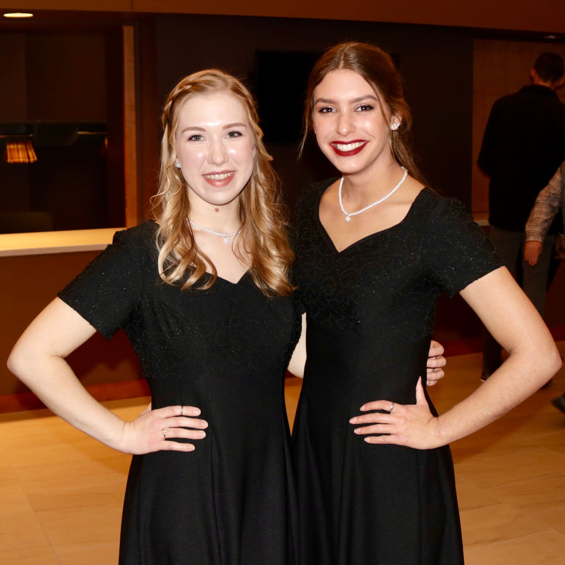 Two women in black dresses pose, smiling, arms on hips. Indoor, neutral background.