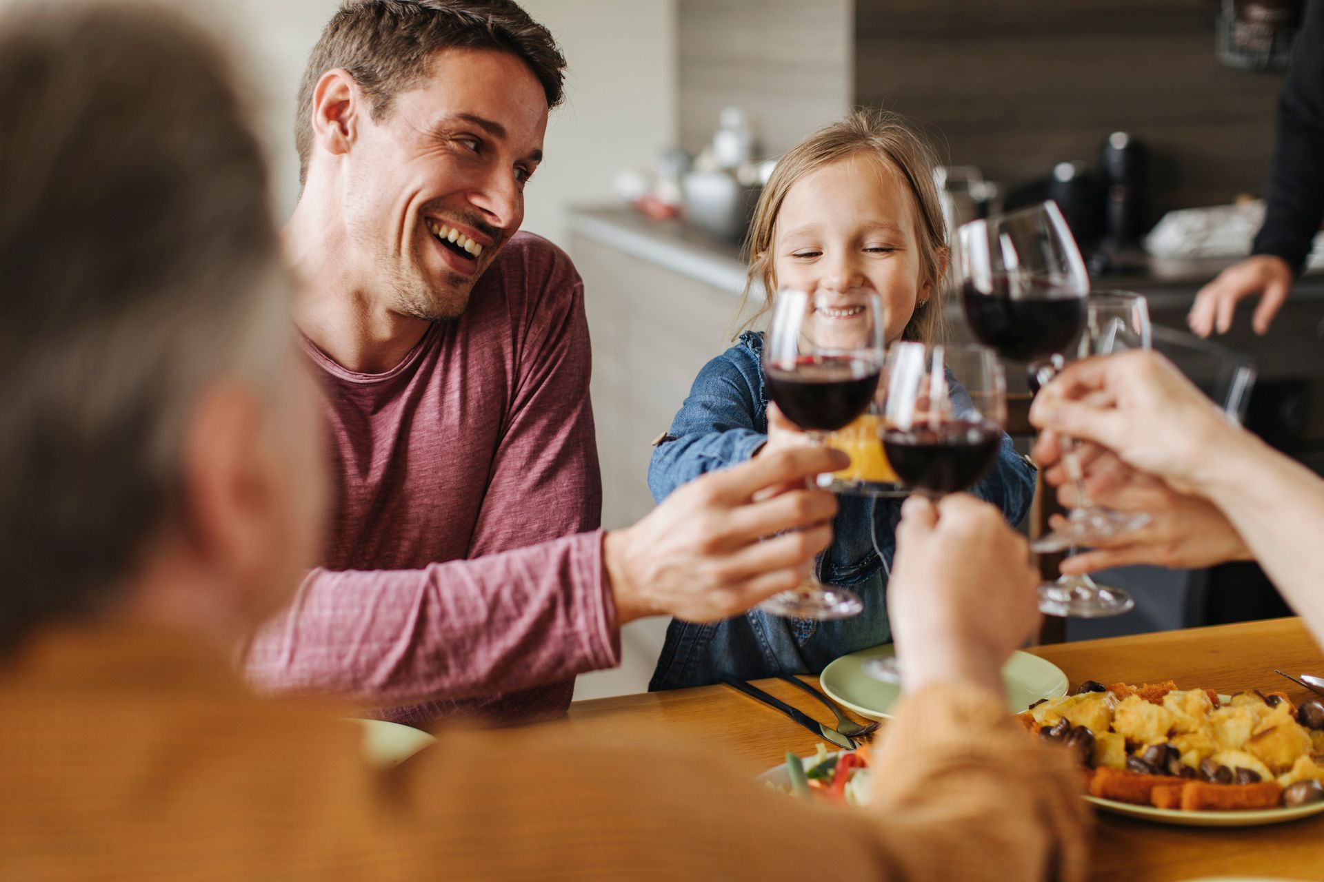 Family clinking glasses of red wine at a dining table. Man laughing, girl smiling, food visible.