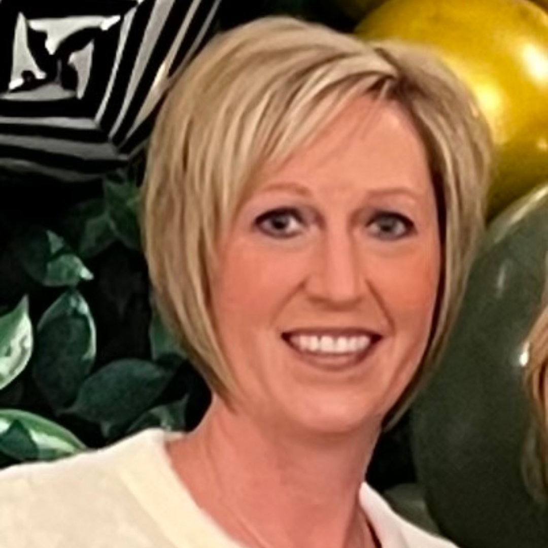 Blonde woman smiling, close-up. Wearing white top. Green and gold balloons in background.