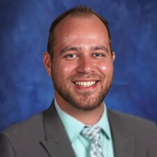 Man smiling, wearing a gray suit, teal shirt, and patterned tie, against a blue background.