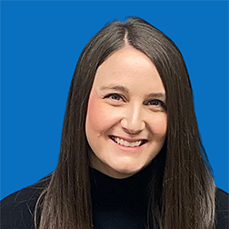 Woman with long brown hair smiling at the camera, wearing a black shirt, against a blue background.