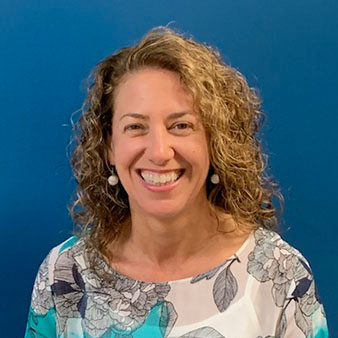 Woman with curly brown hair smiling in front of a blue background. She wears a floral print blouse.