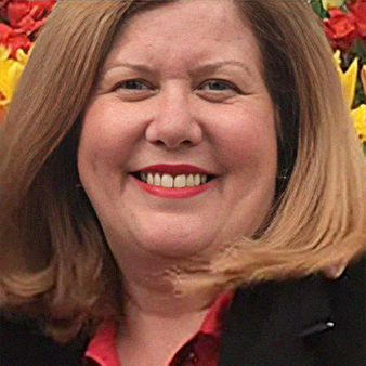 Woman with shoulder-length blonde hair smiling, wearing a red shirt and black blazer, in front of flowers.