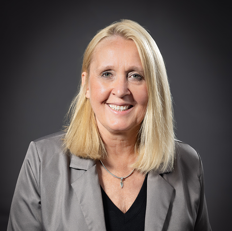Blonde woman in grey blazer smiles, looking at camera. Dark background.