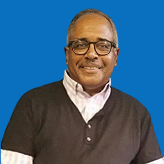 Man with glasses smiles, wearing a black shirt over a white collared shirt, blue background.