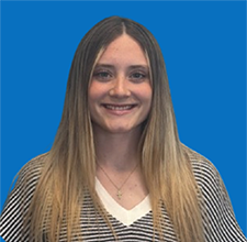Young woman with brown hair and blue eyes smiles at the camera. Blue background.