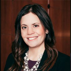 Woman with dark hair smiles, wearing a pearl necklace and blazer, in a wood-paneled room.