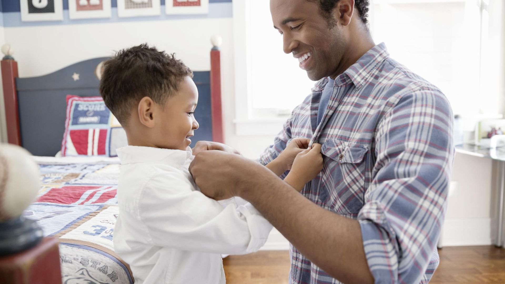 Father buttoning his young son's shirt in a bedroom. Both are smiling.