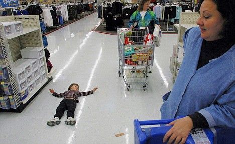 Child lying on floor in store as woman looks on; another shopper with cart in background.