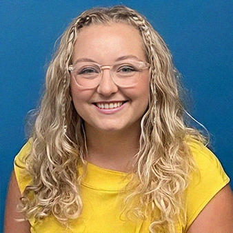 Woman with blonde hair, braids, and glasses smiles, wearing a yellow top against a blue backdrop.