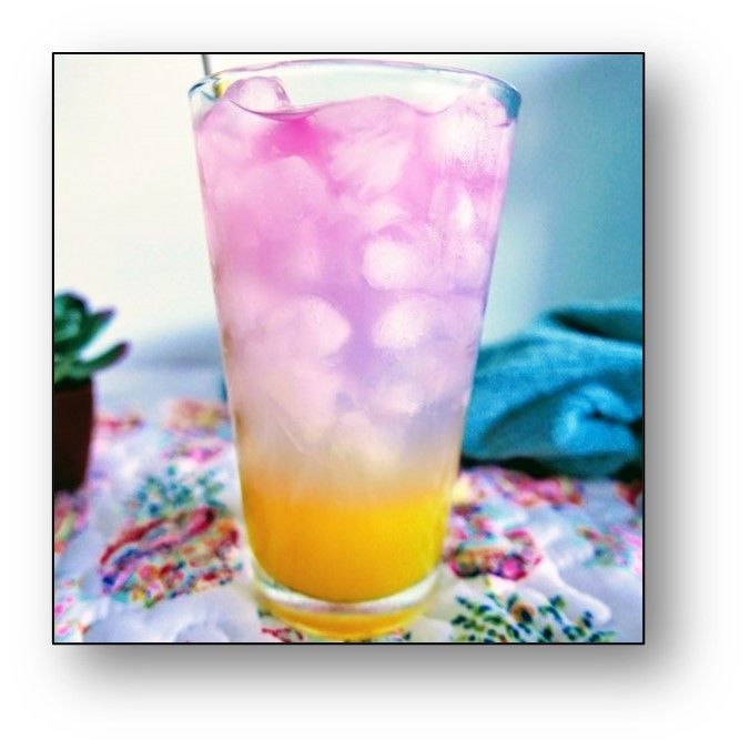 Tall glass with layered yellow and pink drink, filled with ice, on floral tablecloth.