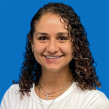Woman with curly brown hair smiling, wearing a white shirt and necklaces against a blue background.