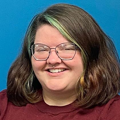 Person with glasses and a nose ring smiles, light green hair highlights against a blue background.