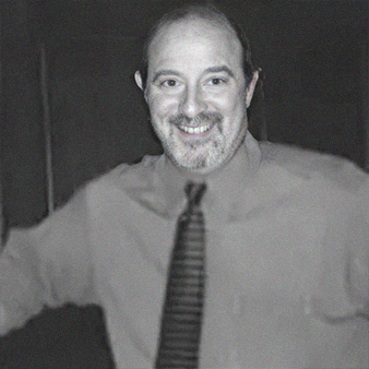 Smiling man in a button-down shirt and tie, arms outstretched, in a neutral setting.