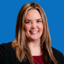 Woman with long blonde hair smiling, wearing a red shirt and black blazer against a blue background.