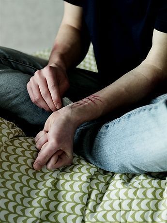 Person sitting on a bed, wearing jeans and a dark shirt, with visible self-inflicted wounds on their wrist.