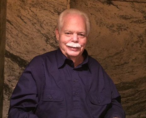Smiling older man in a blue shirt with a white mustache against a textured wall.