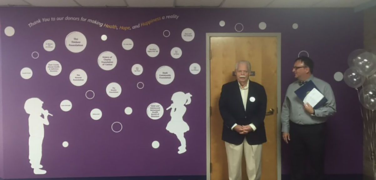 Two men standing in front of a purple wall with children's art; one holds a folder, one is wearing a blazer.