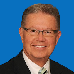 Man with glasses smiling, wearing a black suit and tie against a blue background.