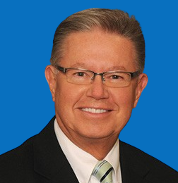 Man in suit and glasses smiles at camera against a blue background.
