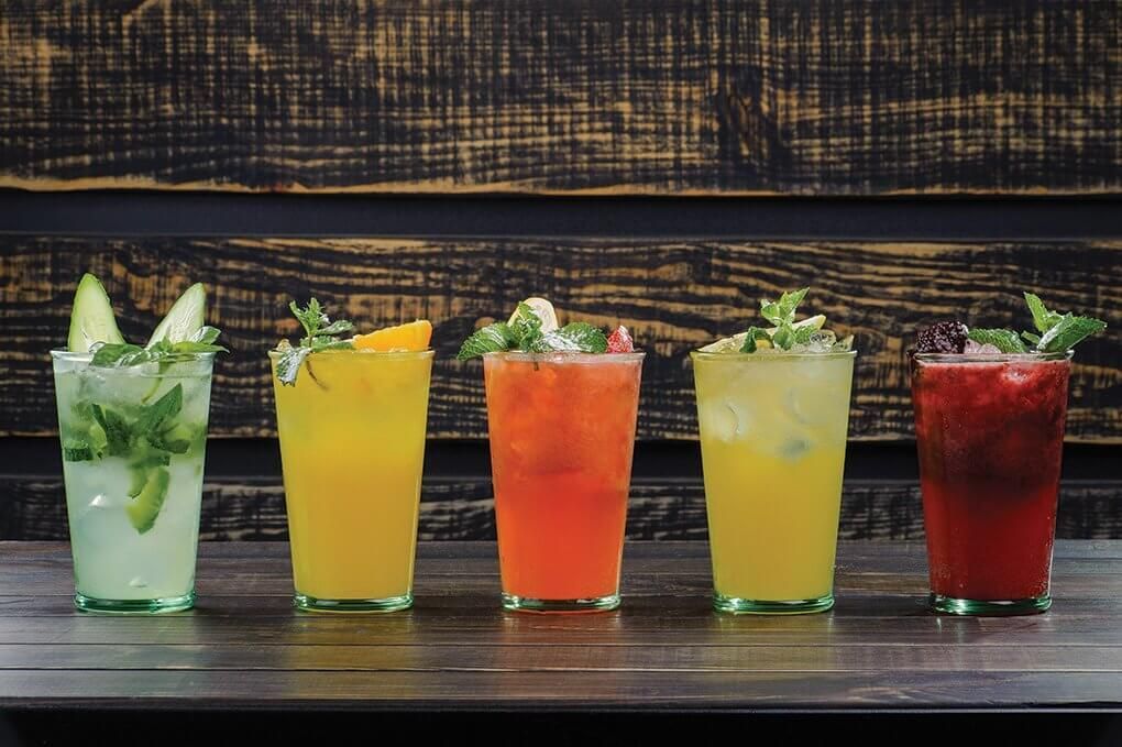 Five colorful cocktails in tall glasses, garnished with fruit and mint, on a wooden bar.