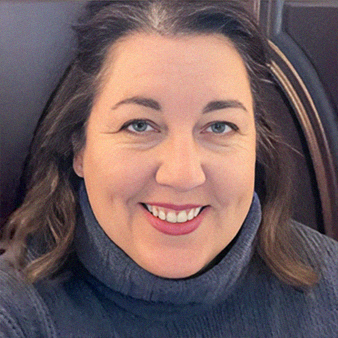 Woman with graying hair smiles at the camera, wearing a blue turtleneck, indoor setting.