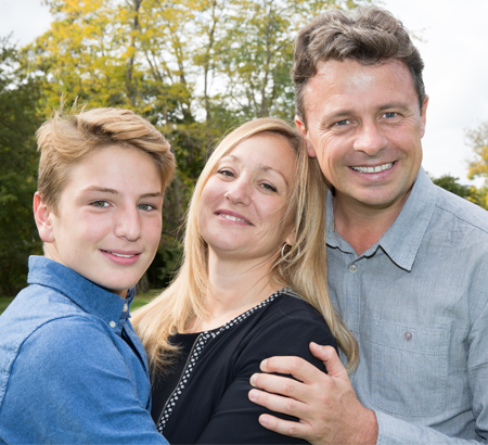 Family of three smiling, posed outdoors: teen son in blue, mother and father with arms around each other.