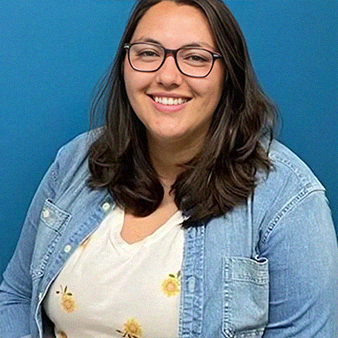 Woman smiling, wearing glasses, denim jacket, floral shirt, against a blue wall.