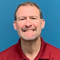 Man with short brown hair and beard smiling, wearing a red shirt against a blue background.