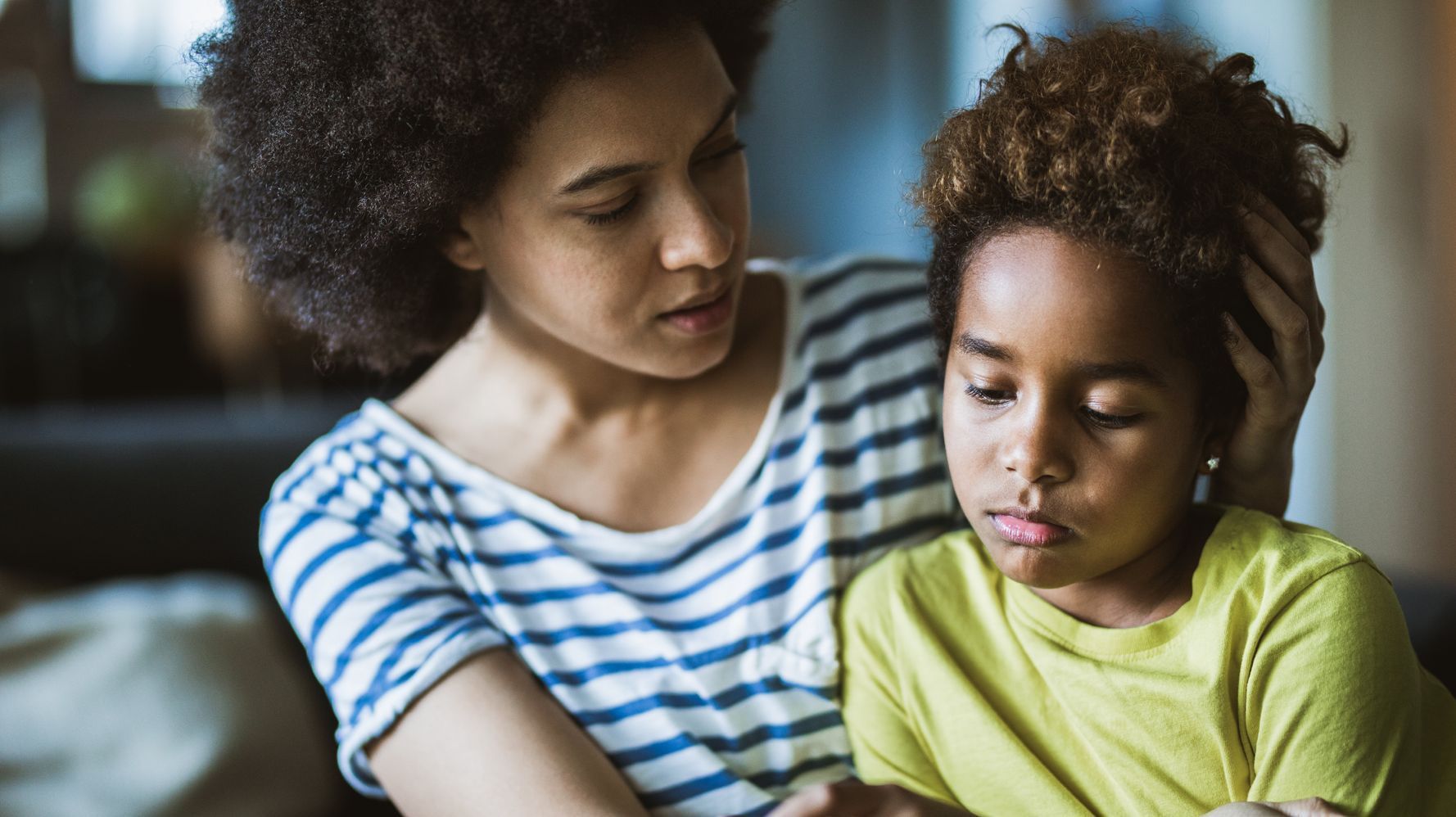 Mother comforts child, hand on head, both looking concerned, indoors.