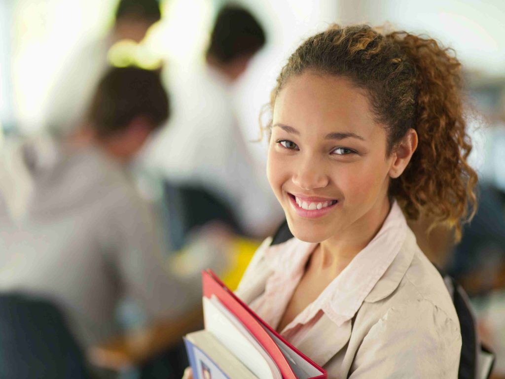 Smiling young woman, holding books, in a classroom, looking at the camera.