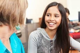 Smiling girl talking to a woman. Girl has long dark hair, gray hoodie. Woman in blue top. Classroom setting.