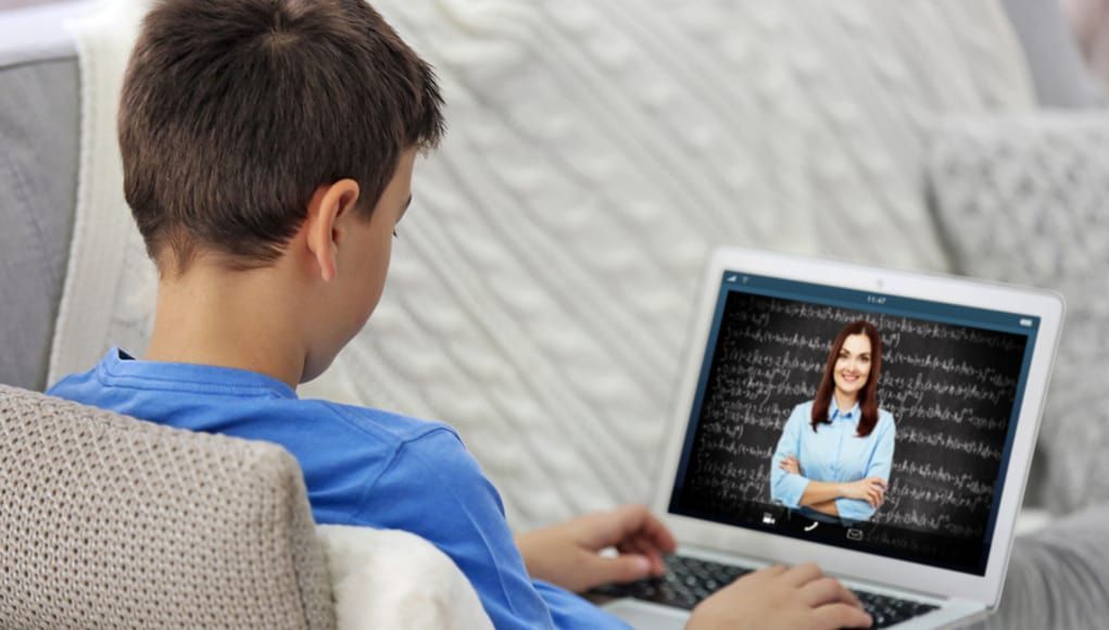 Boy attends virtual class on laptop, sitting on a couch. Teacher on screen, smiles.