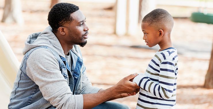 Father and son sitting outdoors, talking. Father holds son's hands, looking concerned. Son looks back.