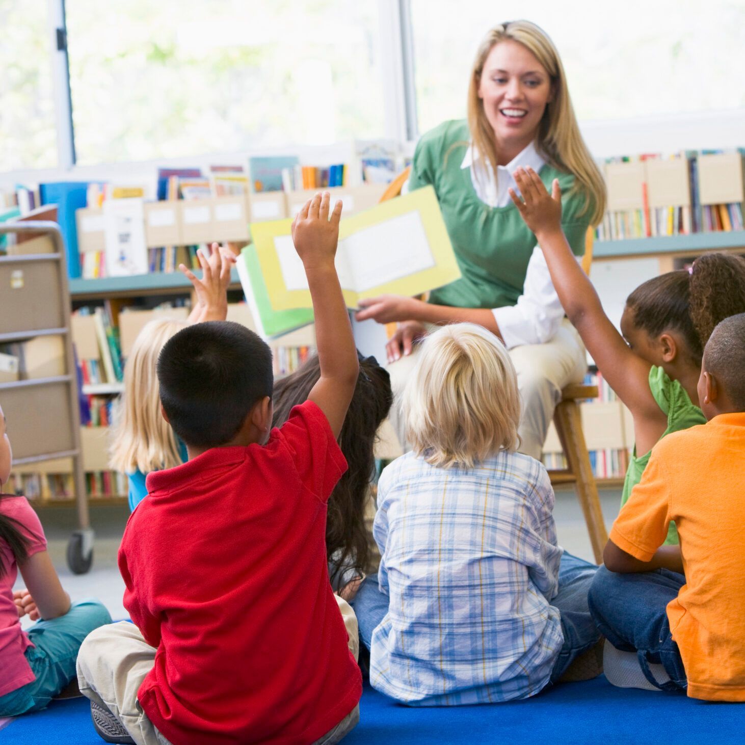 Teacher reading to students in a classroom; children raise hands, smiling; bookshelves in background.