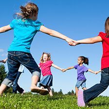 Children in a circle holding hands, jumping on a grassy field under a blue sky.