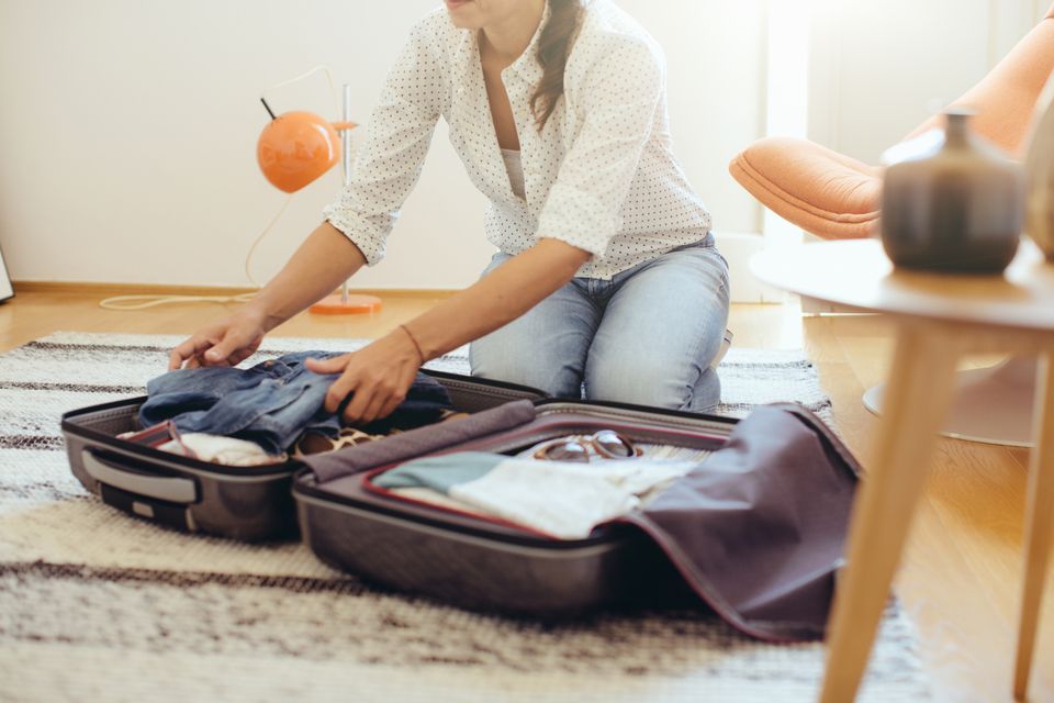 Woman kneeling on a rug, packing a suitcase with clothes. Sunlight streams into the room.